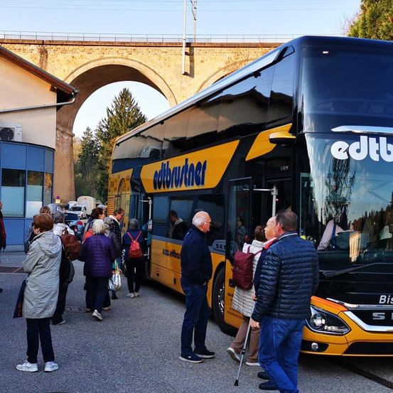 Eine Gruppe von Menschen steht in der Nähe eines gelben und schwarzen Busses, wahrscheinlich wartend, um einzusteigen. Ein Mann benutzt einen Stock, während andere Taschen tragen. Eine Brücke und ein Gebäude sind im Hintergrund.