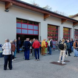 Eine Gruppe von Menschen steht vor einem Gebäude mit großen roten Türen. Sie sind warm gekleidet. Einige tragen Taschen.