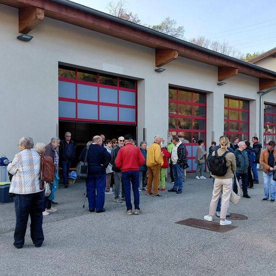 Eine Gruppe von Menschen steht vor einem Gebäude mit großen roten Türen. Sie sind warm gekleidet. Einige tragen Taschen.