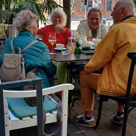 Vier Personen sitzen auf einer Terrasse an einem Tisch. Die Frau in Blau und Grün trägt einen Rucksack. Die Frau in Rot zeigt den Daumen hoch. Die Männer tragen Jacken und Sneakers.