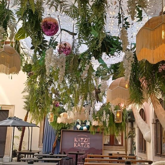 Ein Restaurant mit Tischen, Stühlen und einer bunten Deckendekoration. Ein lila Banner mit dem Wort Katze ist in der Mitte. Ein schwarzer Schirm ist auf der linken Seite. Eine Pflanze und eine Lampe sind auf der rechten Seite.
