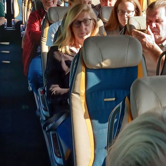 Mehrere Menschen sind in einem Bus, einige sitzen und andere stehen. Eine Frau mit Brille sitzt in der Mitte, während ein Mann neben ihr in sein Handy schaut. Eine andere Person hinten trägt eine rote Jacke.