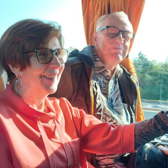 Ein älteres Paar mit Brille sitzt lächelnd in einem Bus. Die Frau trägt ein rotes Blusenhemd, der Mann einen braunen Mantel. Dahinter ist ein Vorhang, und draußen gibt es Bäume und eine Straße.