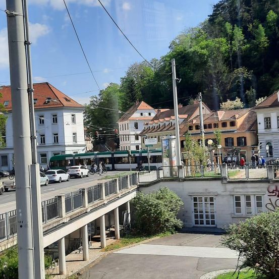 Eine Straßenbahn überquert eine Brücke in einer Stadt mit vielen Gebäuden und Bäumen. Autos und Fahrräder befinden sich auf der Straße darunter.