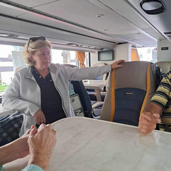 Eine Frau mit Sonnenbrille steht in einem Bus, zeigt auf einen Sitz und zwei Personen halten einen Tisch vor ihr. Der Bus hat einen Monitor an der Wand befestigt.
