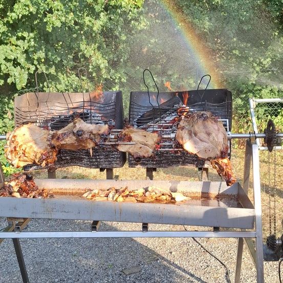Zwei große Fleischstücke werden im Freien auf einem Grill geröstet, mit einem Regenbogen im Hintergrund.