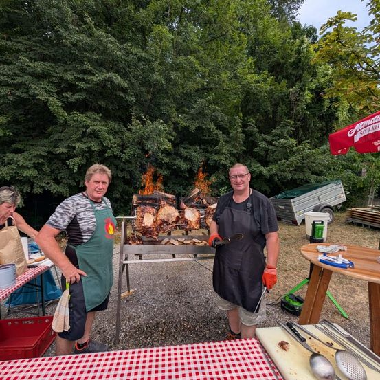 Zwei Männer in Schürzen stehen neben einem Grill mit kochendem Fleisch. Eine Frau steht hinter ihnen, und ein Tisch mit Utensilien steht vor ihnen.