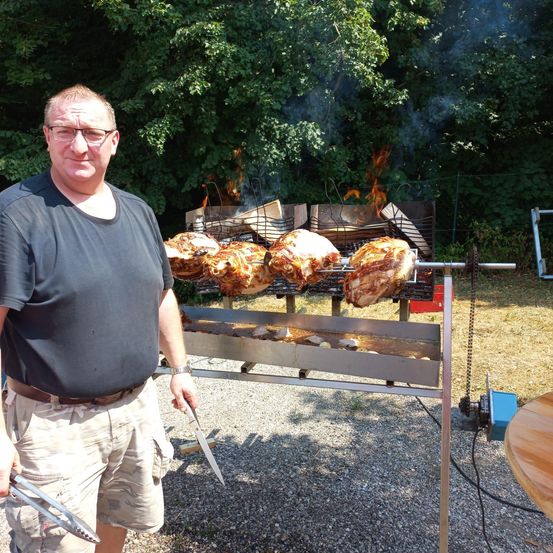 Ein Mann steht neben einem Grill mit mehreren gebratenen Hähnchen darauf, hält Zange und Messer in der Hand.
