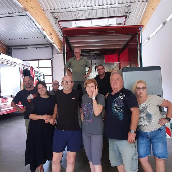 Eine Gruppe von Menschen steht in einer Garage mit einem Feuerwehrauto im Hintergrund. Sie lächeln und scheinen für ein Foto zu posieren.