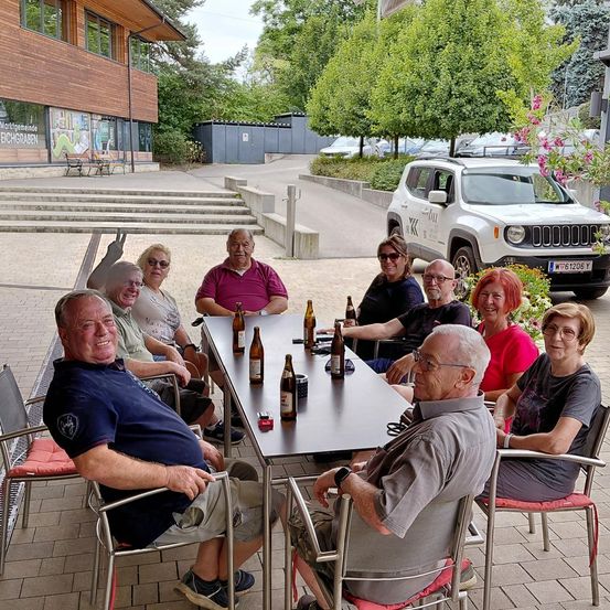 Eine Gruppe älterer Menschen sitzt draußen um einen Tisch, mit Flaschen und einem Jeep im Hintergrund.