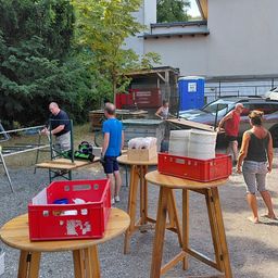 Eine Gruppe von Menschen richtet Tische mit roten Kisten und Tellern für eine Outdoor-Veranstaltung in einem Kiesbereich ein. Bäume und ein Gebäude sind im Hintergrund.