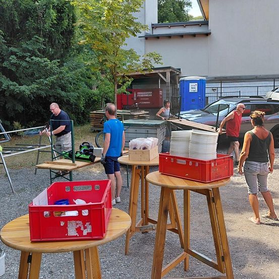 Eine Gruppe von Menschen richtet Tische mit roten Kisten und Tellern für eine Outdoor-Veranstaltung in einem Kiesbereich ein. Bäume und ein Gebäude sind im Hintergrund.