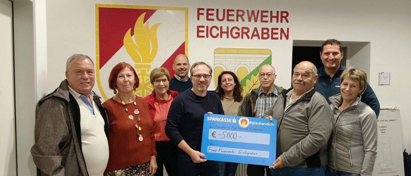 Eine Gruppe von Menschen steht vor einer Wand mit dem Text 'Feuerwehr Eichgraben' und einem Logo. Ein Mann hält einen blauen Scheck über 5000 Euro von den Freunden der Eichgrabener Feuerwehr.