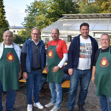 Fünf Männer in Schürzen posieren für ein Foto vor einem Gewächshaus mit Bäumen im Hintergrund.