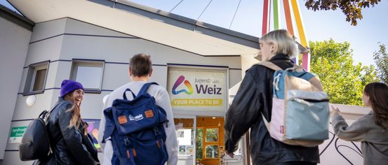Drei Personen mit Rucksäcken gehen auf ein Gebäude zu, an dem ein Schild mit der Aufschrift Jugendhaus Welz steht.