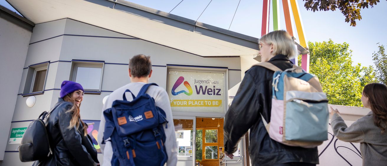 Drei Personen mit Rucksäcken gehen auf ein Gebäude zu, an dem ein Schild mit der Aufschrift Jugendhaus Welz steht.