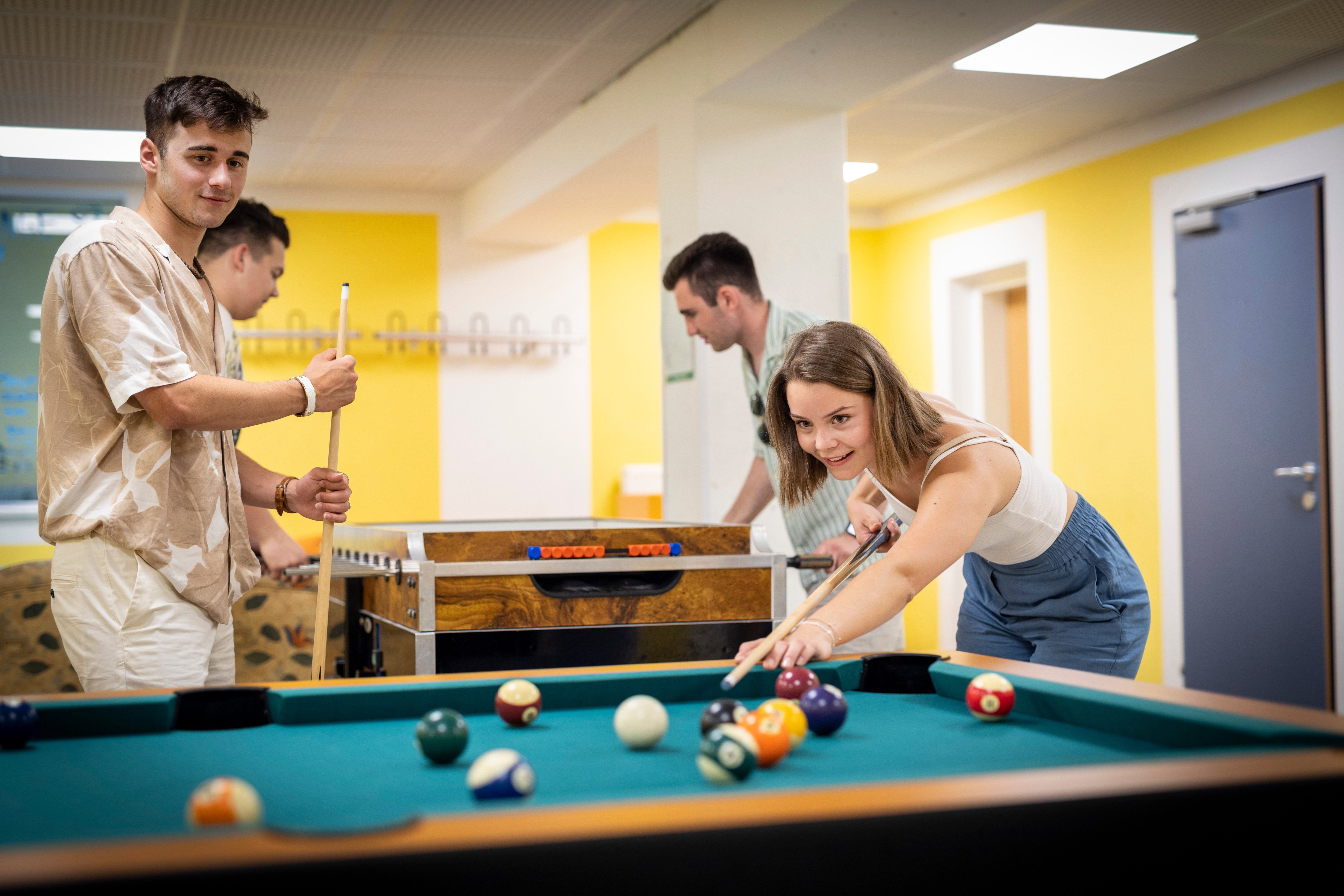Drei junge Erwachsene spielen Billard in einem hell erleuchteten Raum. Die Frau beugt sich vor und zielt auf die Bälle. Die beiden Männer beobachten.