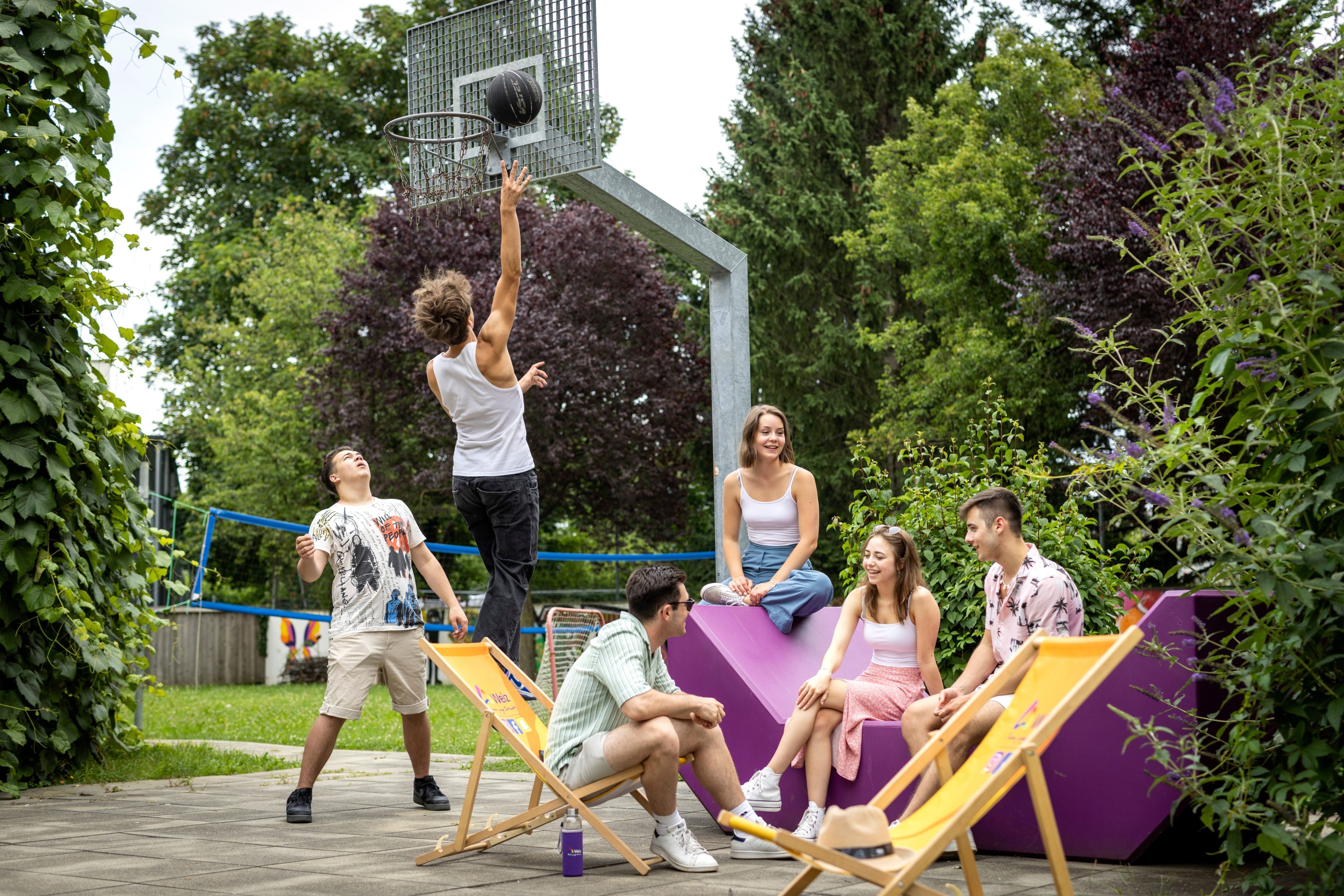 Eine Gruppe von Freunden spielt Basketball im Freien. Eine Person springt, um einen Korb zu werfen, während andere auf violetten Stühlen sitzen und zuschauen.