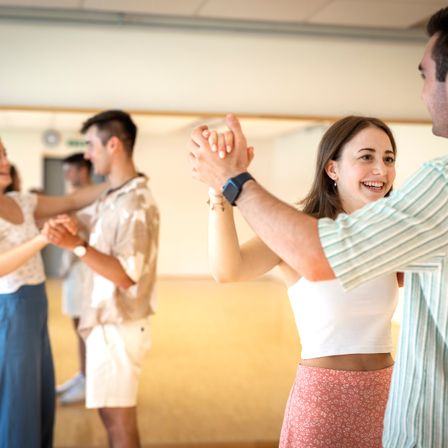 Eine Gruppe von Menschen tanzt in einem Studio. Eine Frau lächelt, während ein Mann ihre Hände hält. Dahinter tanzt ein weiteres Paar. Das Studio hat Holzböden und weiße Wände.