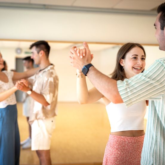 Eine Gruppe von Menschen tanzt in einem Studio. Eine Frau lächelt, während ein Mann ihre Hände hält. Dahinter tanzt ein weiteres Paar. Das Studio hat Holzböden und weiße Wände.
