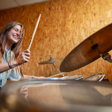 Eine junge Frau lächelt, während sie in einem Raum mit Holzwänden Schlagzeug spielt. Sie hält einen Drumstick und scheint sich zu amüsieren.