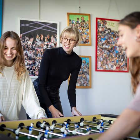 Drei Frauen lächeln und schauen auf ein Tischfußballspiel. Eine Frau trägt eine Brille und ein schwarzes Rollkragenshirt. Hinter ihnen hängen drei Bilder an der Wand.