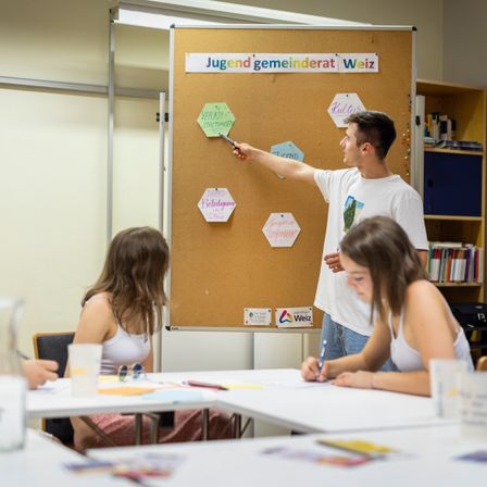 Ein Mann zeigt auf eine Tafel mit verschiedenen farbigen Sechsecken mit Themen wie 'Veraint Staeltungen'. Zwei Frauen sitzen mit Stiften und Papier an einem Tisch.