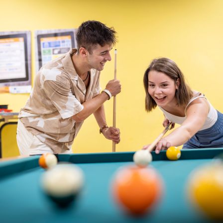 Ein Mann und eine Frau spielen Billard in einem hell erleuchteten Raum, lächelnd und engagiert. Der Mann hält einen Billardstock, und die Frau zielt auf die Bälle. Der Billardtisch ist mit bunten Kugeln gefüllt.