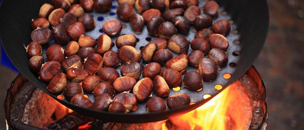 Eine Bratpfanne voller gerösteter Kastanien liegt über einem offenen Feuer. Die Kastanien sind braun und golden, einige noch in ihrer Schale.