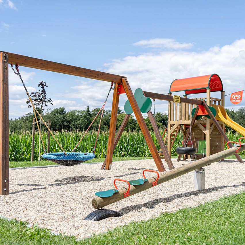 Ein Spielplatz mit Schaukeln, einer Rutsche und einem Reifenschaukel. Es gibt eine gelbe Rutsche und ein rotes Dach. Bäume und Maisfelder sind im Hintergrund.