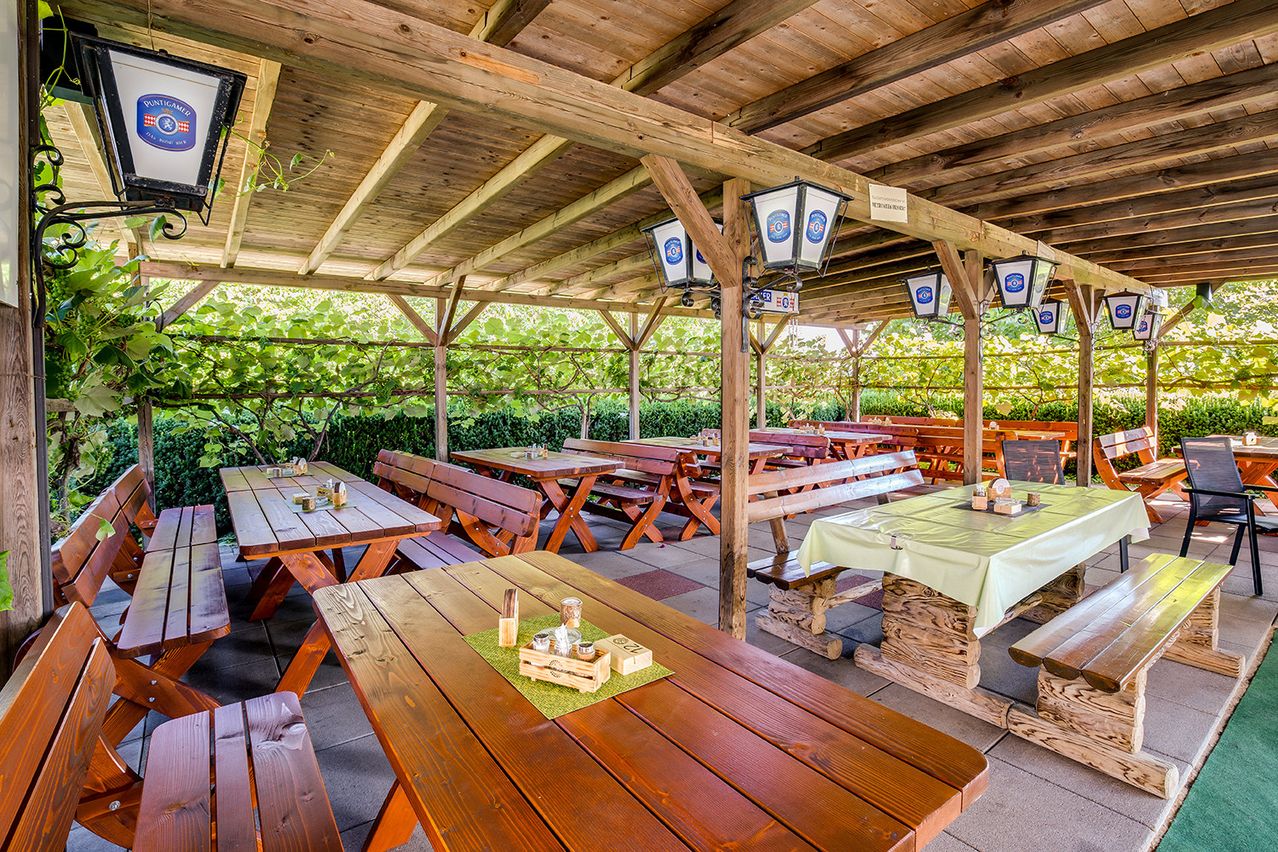 Ein Freiluftrestaurant mit Holztischen und Bänken unter einem hölzernen Vordach, umgeben von Grün. Laternen hängen von der Decke.
