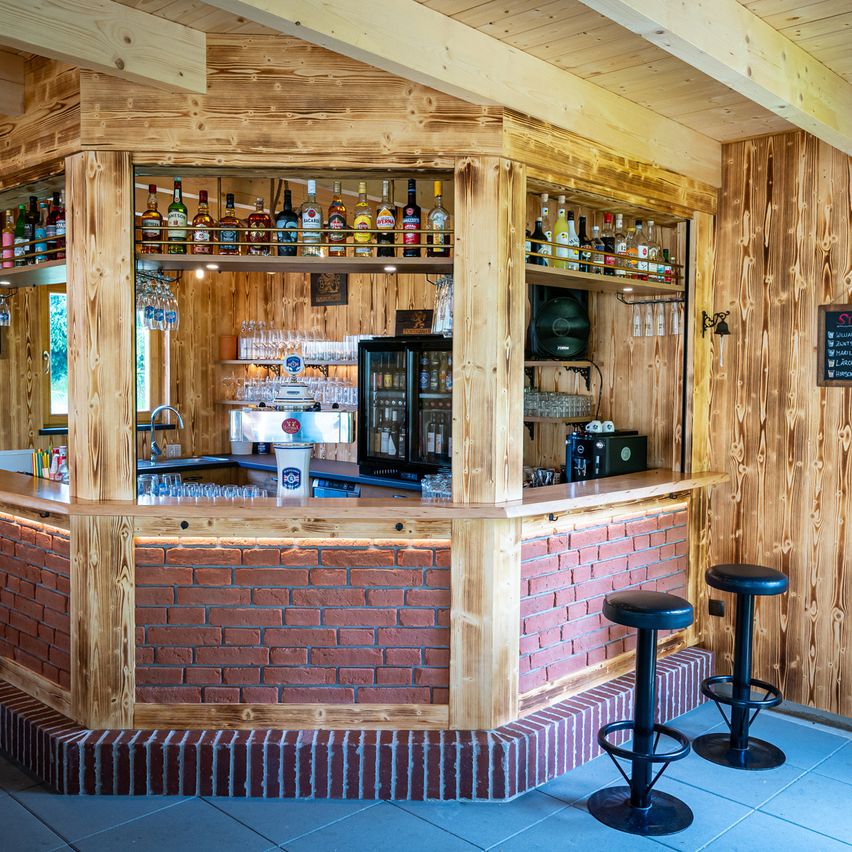 Eine rustikale Bar mit Holzregalen, die verschiedene Likörflaschen halten, einer Ziegelwand und zwei schwarzen Stühlen. Im Hintergrund befinden sich ein Kühlschrank, ein Waschbecken und eine Tafel mit Speisekarte.