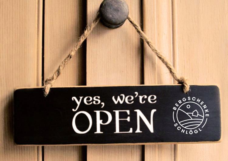 Ein schwarzes Schild mit weißem Text, der 'ja, wir sind offen' sagt, ist an einer Holztür mit einem Seil befestigt. Ein Logo befindet sich auf der rechten Seite des Schildes.