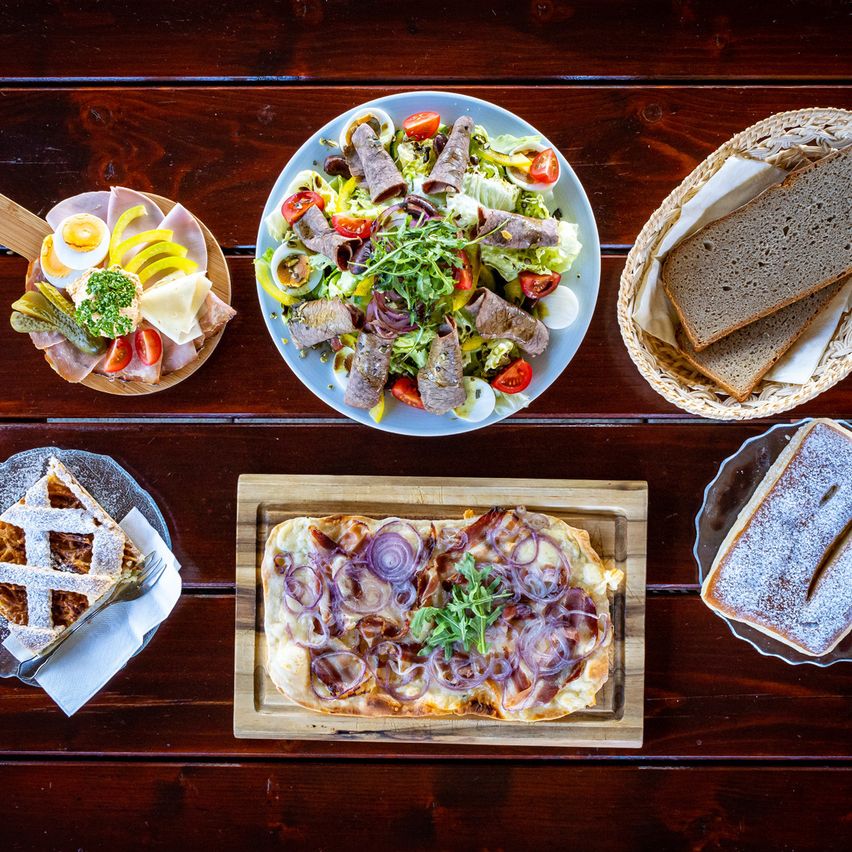 Auf einem Holztisch sind verschiedene Speisen zu sehen: ein Salat mit Fleisch, Eiern und Gemüse; ein Käseteller; Brot in einem Korb; eine Pizza mit Zwiebeln und Fleisch; und ein Dessert.