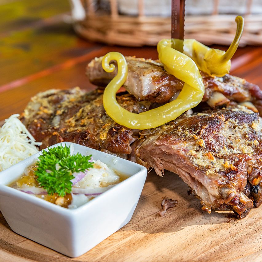 Auf einem Holzteller liegen gegrillte Rippchen mit gelben Paprikaschoten, ein weißes Schälchen mit Petersilie, Zwiebeln und Dip steht daneben.