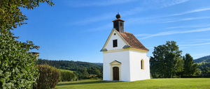 Eine kleine weiße Kapelle mit einem braunen Dach und einem schwarzen Turm steht auf einem grasbewachsenen Hügel unter einem klaren blauen Himmel.