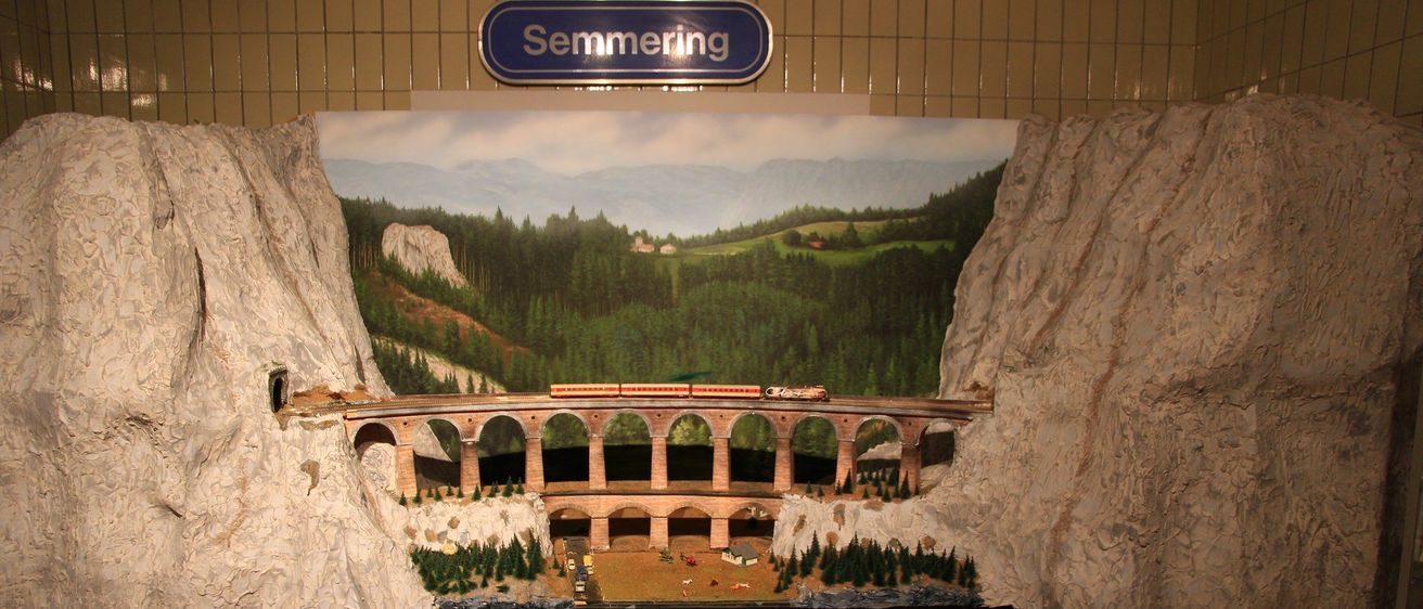 Eine Modelleisenbahnszene zeigt einen Zug, der eine Brücke über einen Berg überquert. Darüber steht ein Schild mit der Aufschrift 'Semmering'. Unter der Szene befindet sich ein Warnschild mit der Aufschrift 'Das Überschreiten der Gleise ist streng verboten.'