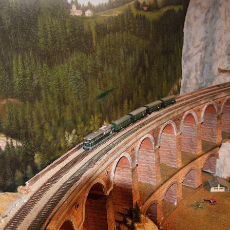Ein Miniaturmodell eines grünen Zuges, der sich auf einer hölzernen Brücke mit Bogenstrukturen bewegt, vor dem Hintergrund dichter Bäume und Berge.