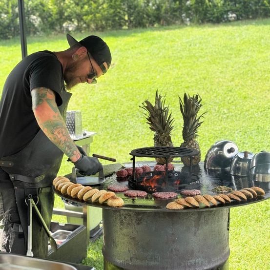 Ein Mann kocht Fleisch auf einem Grill im Freien. Er trägt ein schwarzes Shirt und eine Kappe und hat Tattoos an den Armen. Es gibt Ananas und andere Lebensmittel auf dem Grill.