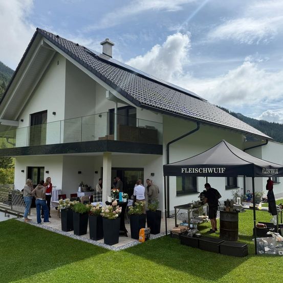 Ein modernes Haus mit Solarpanelen auf dem Dach. Ein Zelt mit Vordach ist im Garten aufgestellt. Menschen sind versammelt, möglicherweise zur Vorbereitung eines Events. Topfpflanzen und Blumen stehen auf dem Boden.