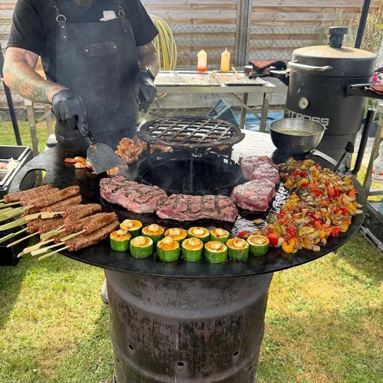 Eine Person in einer Schürze grillt verschiedene Fleischsorten und Gemüse auf einem großen Grill im Freien. Der Grill hat eine Grillrost und eine Schüssel. Ein Tisch mit Gewürzen ist in der Nähe.