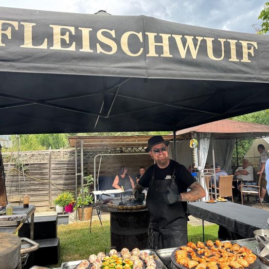 Ein Koch steht unter einem Vordach mit der Aufschrift 'Fleischwuif' und brät Essen. Hinter ihm sind Tische mit Essen und sitzenden Menschen zu sehen. Im Hintergrund befinden sich ein Zaun, Pflanzen und ein Gebäude.