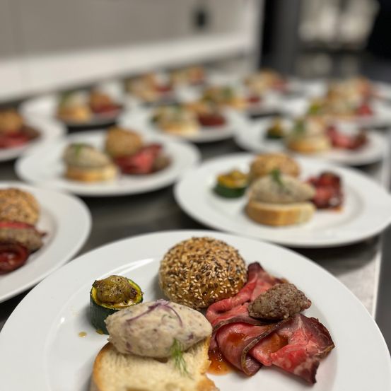 Ein Tisch mit mehreren Tellern verschiedener Gerichte, einschließlich Fleisch, Gemüse und Brot. Im Vordergrund befindet sich ein Teller mit einem Fleischstück, einem Brotstück und einer Gurke.