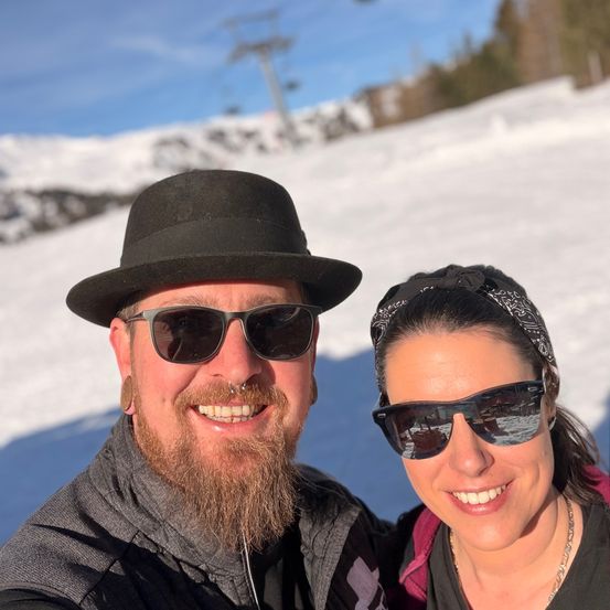 Ein Mann und eine Frau lächeln in die Kamera auf einem verschneiten Berg mit einer Skilift im Hintergrund.