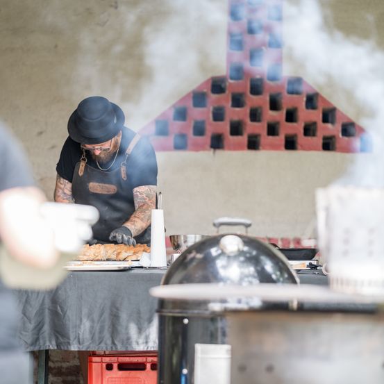 Ein Mann in Hut und Schürze kocht Essen auf dem Grill. Es gibt einen verschwommenen Blick auf eine andere Person.