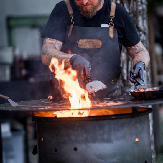 Ein bärtiger Mann kocht mit Feuer auf einem Grill, trägt eine schwarze Schürze und Handschuhe.
