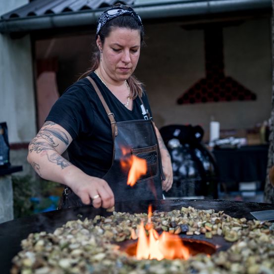 Eine Frau in einer schwarzen Schürze kocht Essen auf einem großen Outdoor-Grill mit Flammen, in einer Außenküche stehend.