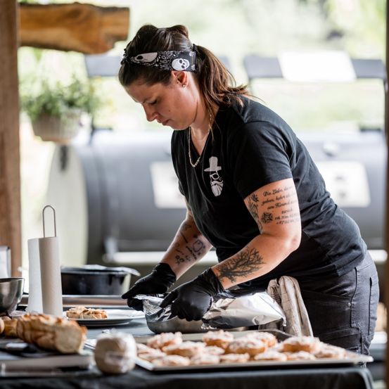 Eine Frau in schwarzen Handschuhen und einem schwarzen Shirt bereitet in einer Outdoor-Küche Essen zu. Sie trägt ein Bandana und hat Tattoos an den Armen.