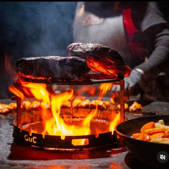 Ein Koch grillt Fleisch in einem schwarzen Metallkorb über offenem Feuer. Ein Gemüseschüssel befindet sich in der Nähe.