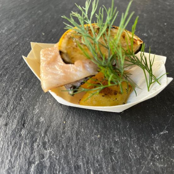 Ein kleines Gericht mit gegrilltem Lachs, Zitrone und Kräutern auf einer Schieferoberfläche. Der Lachs hat eine angekohlte Oberseite und ist mit Zitrone und Kräutern garniert.
